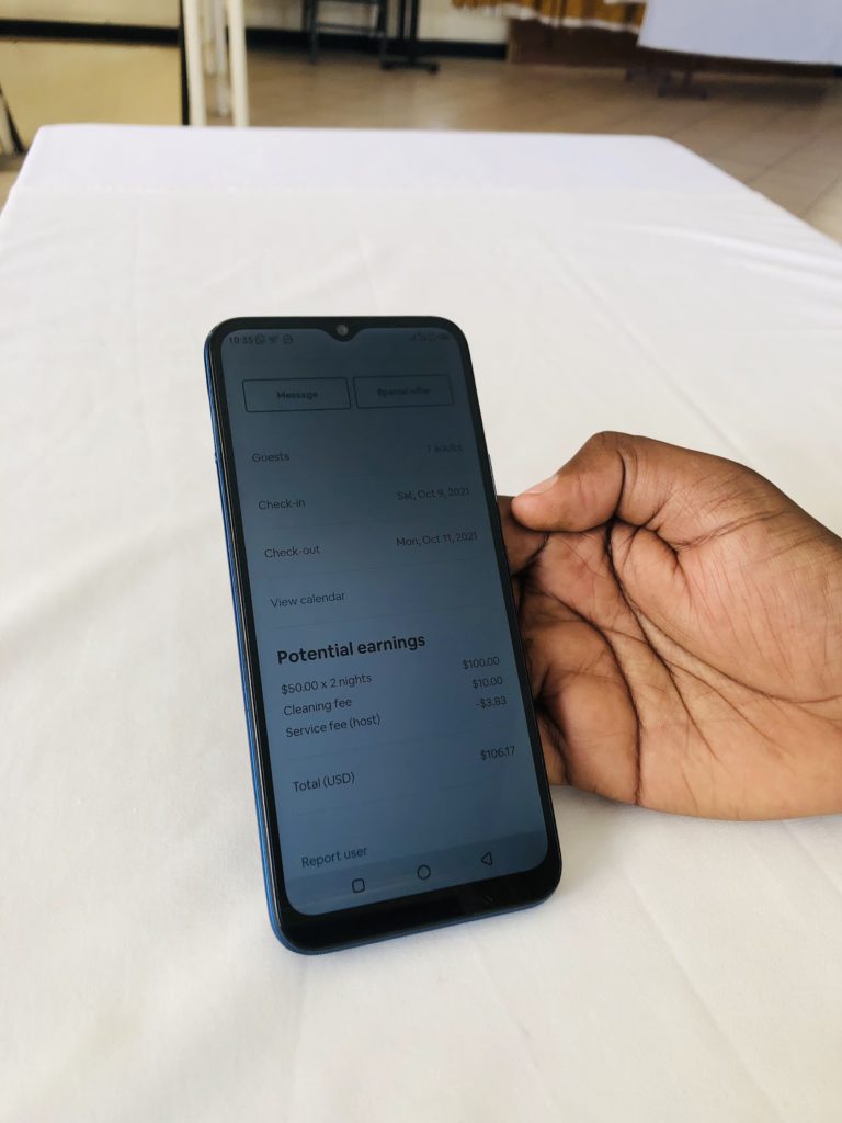 A close-up of someone's hand holding a smartphone, which shows the earnings page on the Airbnb app. The screen shows potential earnings of $106.17 for a two-night stay.