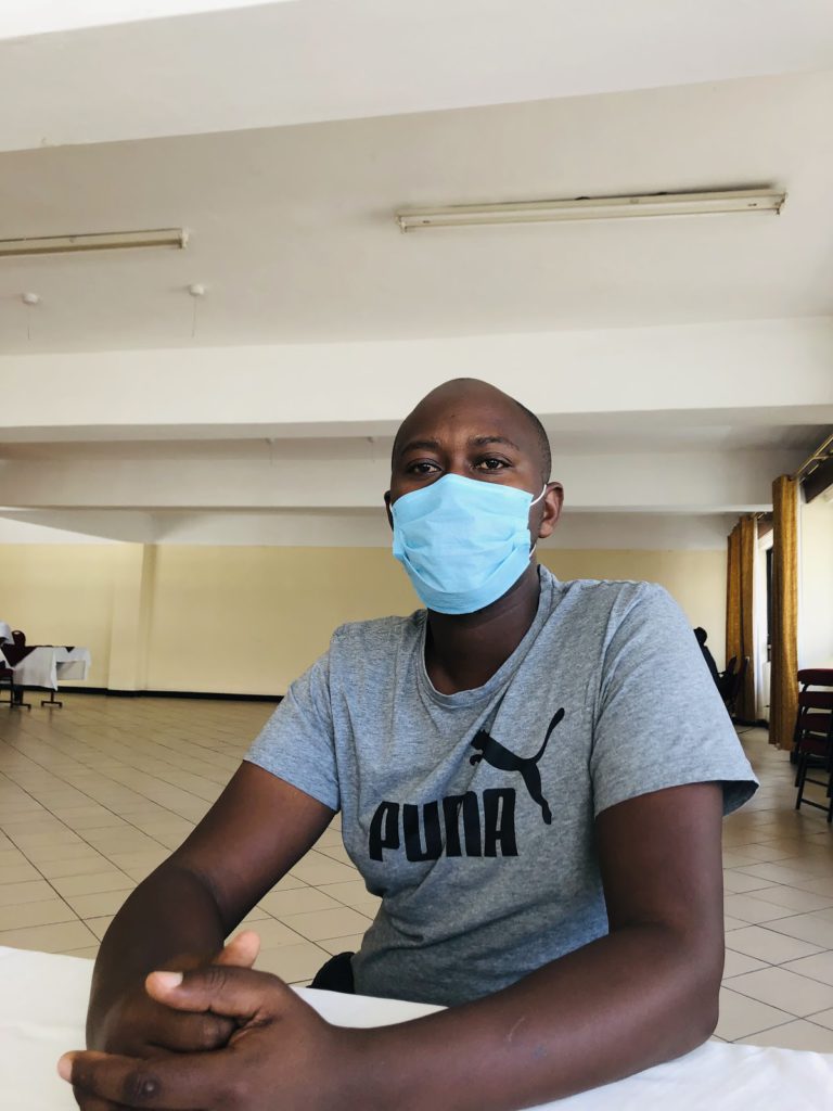 A man poses for a photo in a room after the conversation with the researcher. He is seated at a table and wearing a gray t-shirt with the Puma logo in black, and a mask.