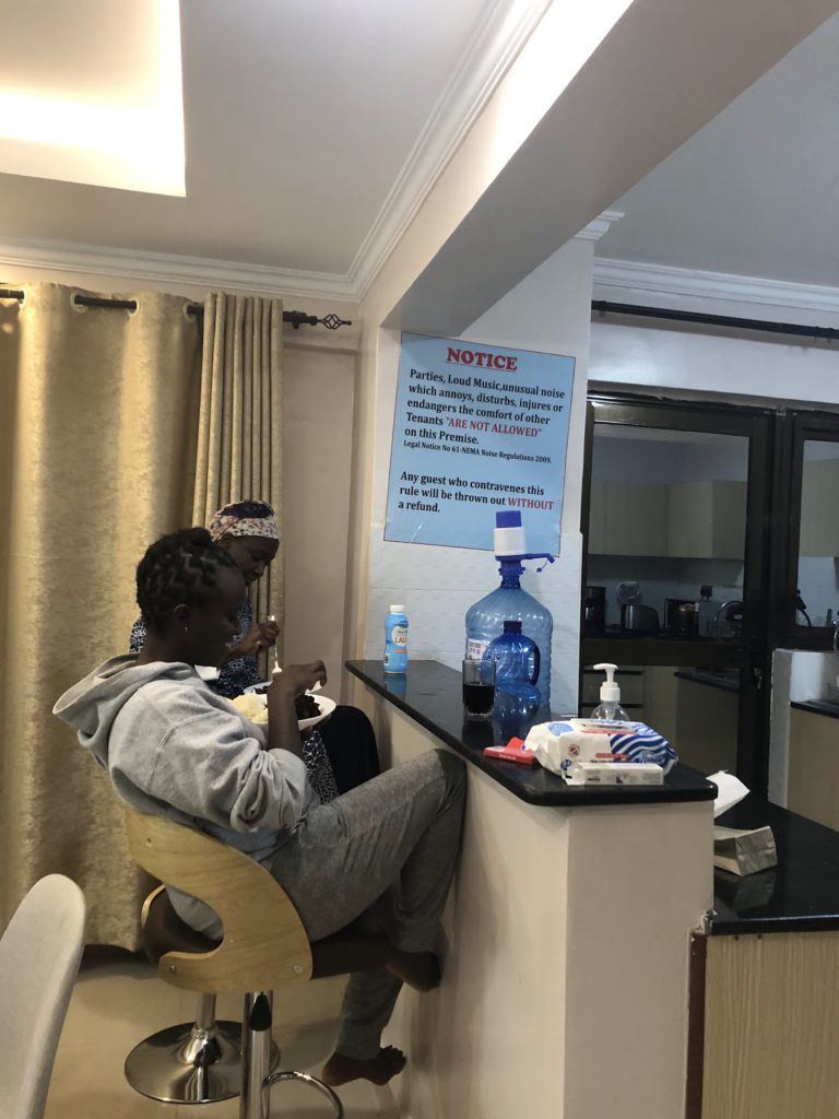 Two women having a meal in an Airbnb with a huge notice on the wall restricting playing of loud music and noise that interferes with other tenants.