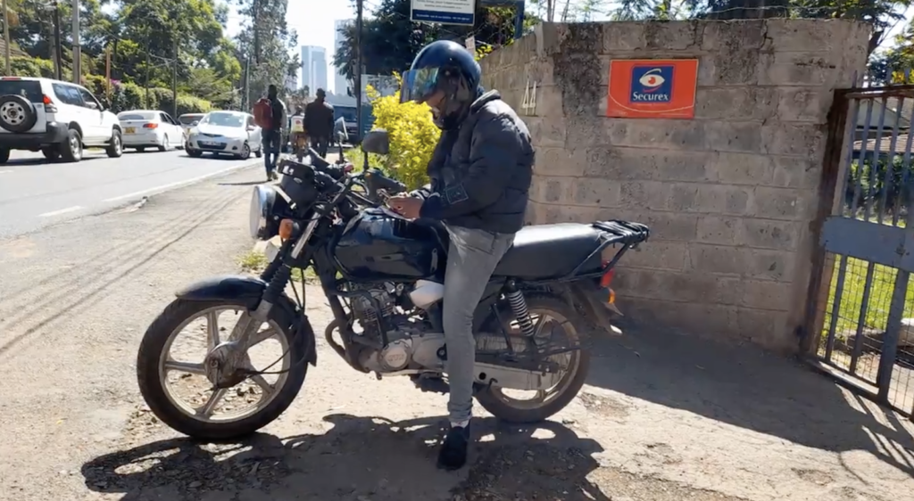 A delivery rider seated on her motorbike with one foot on the ground. She is wearing gray pants, a black puffy jacket, and a black helmet that covers her face. She is looking at her phone.