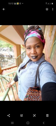 A selfie of a woman. She is standing on a balcony. She is wearing a dark long-sleeved shirt with a light shirt on top and a colorful headscarf. She has dark lipstick.