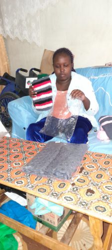 A woman, seated on a couch, packing crochet items in bags. She is wearing blue pants, an orange shirt, and a white jacket. She has long braids. There is a wheelchair next to her.